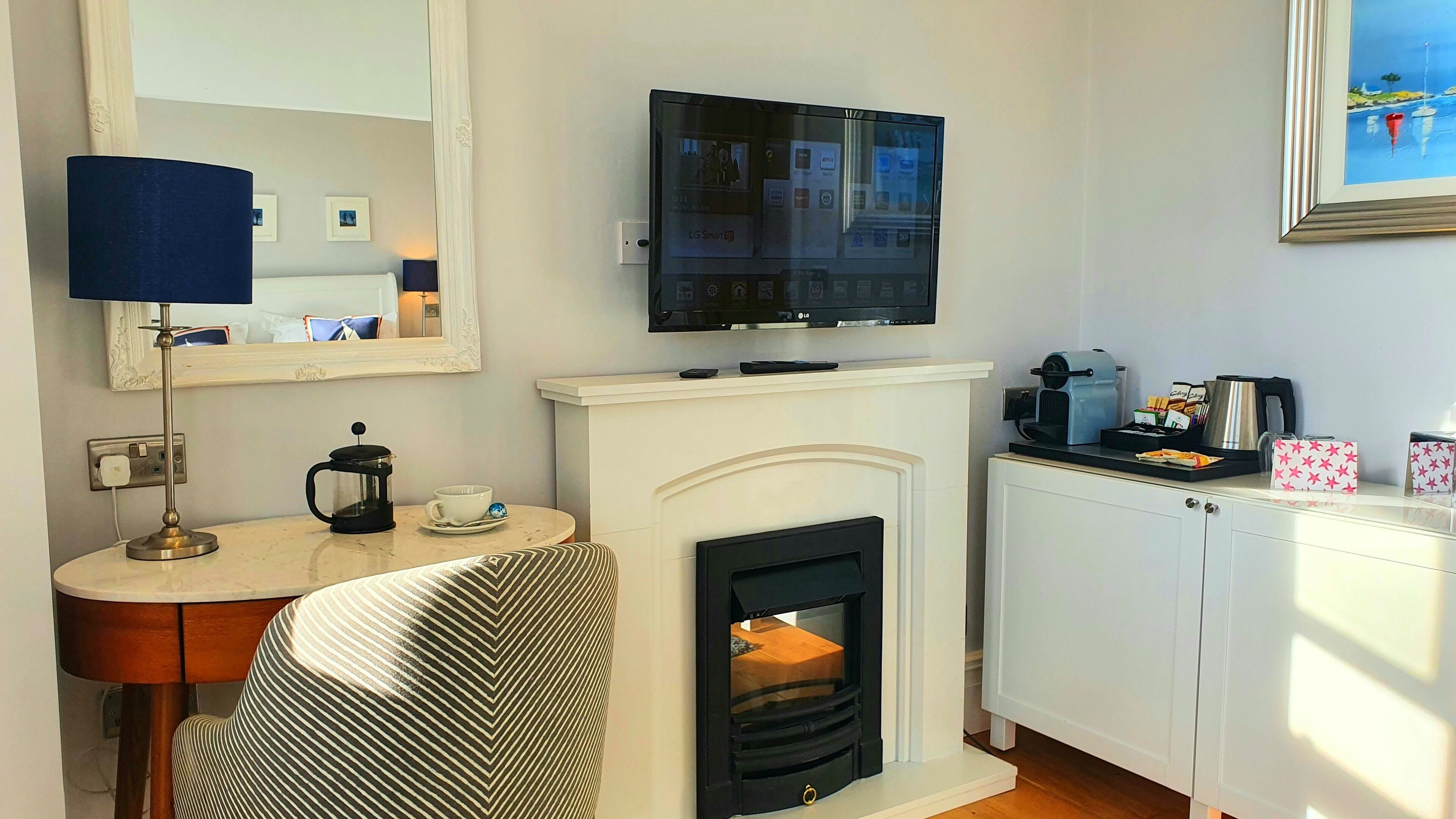 A hotel room with a flat-screen TV mounted above a white fireplace, a coffee machine, and a small table with a lamp and a cup.