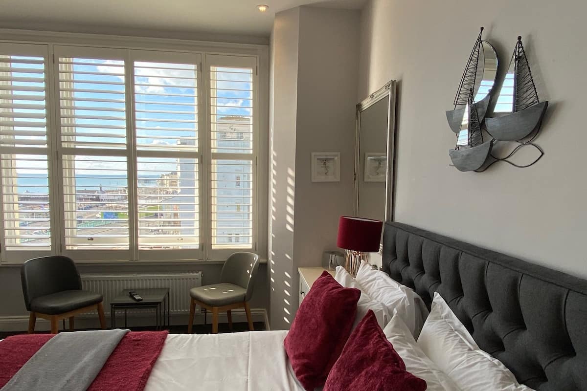 A hotel room with a large bed, two chairs, a desk, a lamp, and a view of the city through large windows.