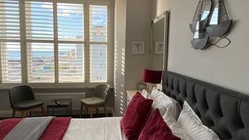 A hotel room with a large bed, two chairs, a desk, a lamp, and a view of the city through large windows.