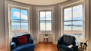 Habitación de esquina con tres ventanas amplias que dan a una playa y un muelle. Hay dos sillones azules y una mesita con una planta.