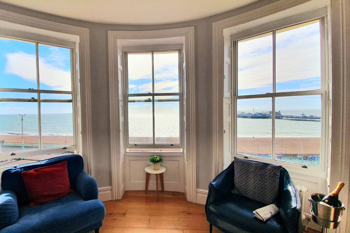 A corner room with three large windows overlooking a beach and a pier. There are two blue armchairs and a small table with a plant.