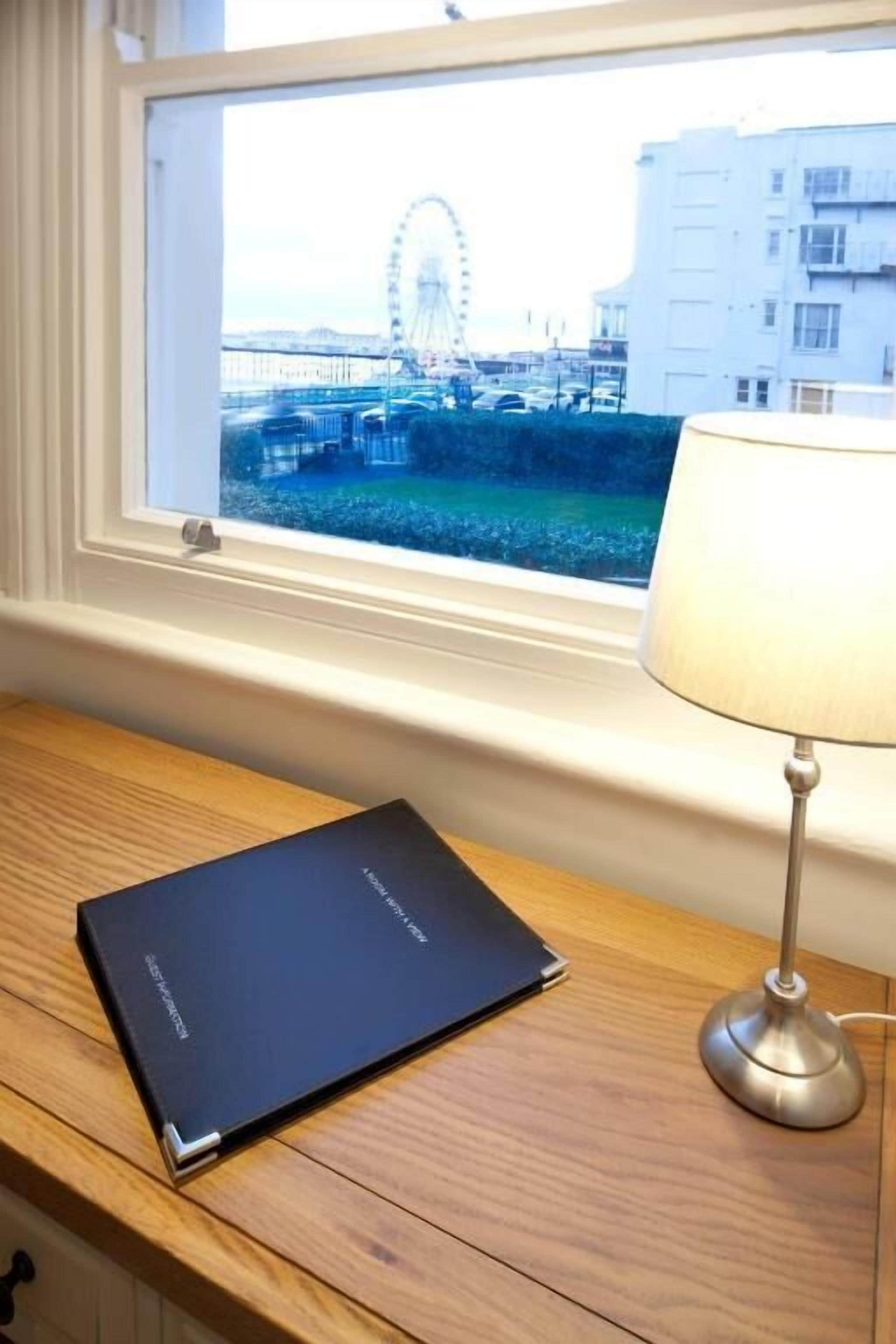 A desk with a book and a lamp by a window overlooking a Ferris wheel and buildings.
