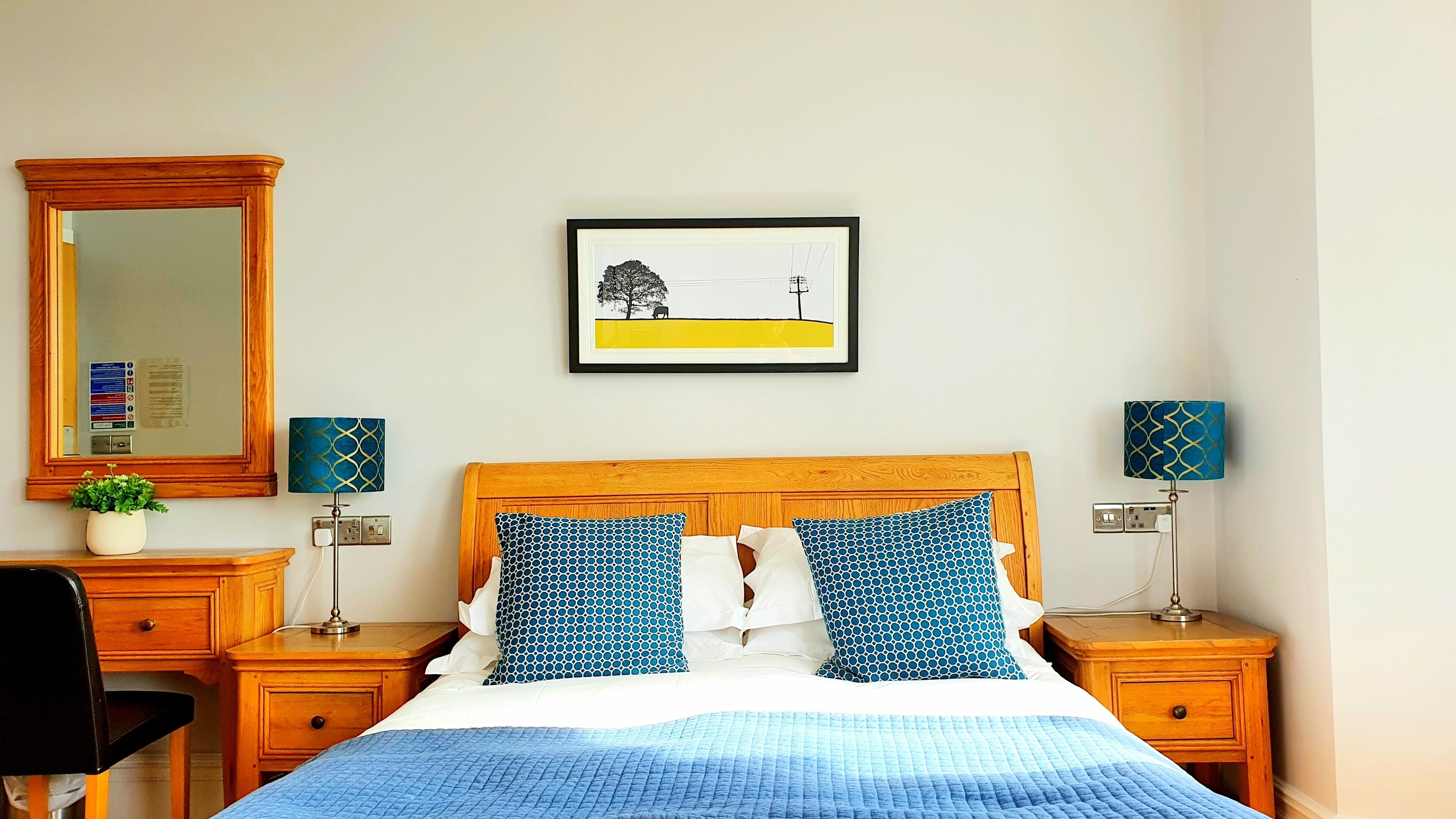 A neatly arranged bedroom with a wooden headboard, a bed with blue bedding, two bedside tables with lamps, a mirror, and a framed picture on the wall.
