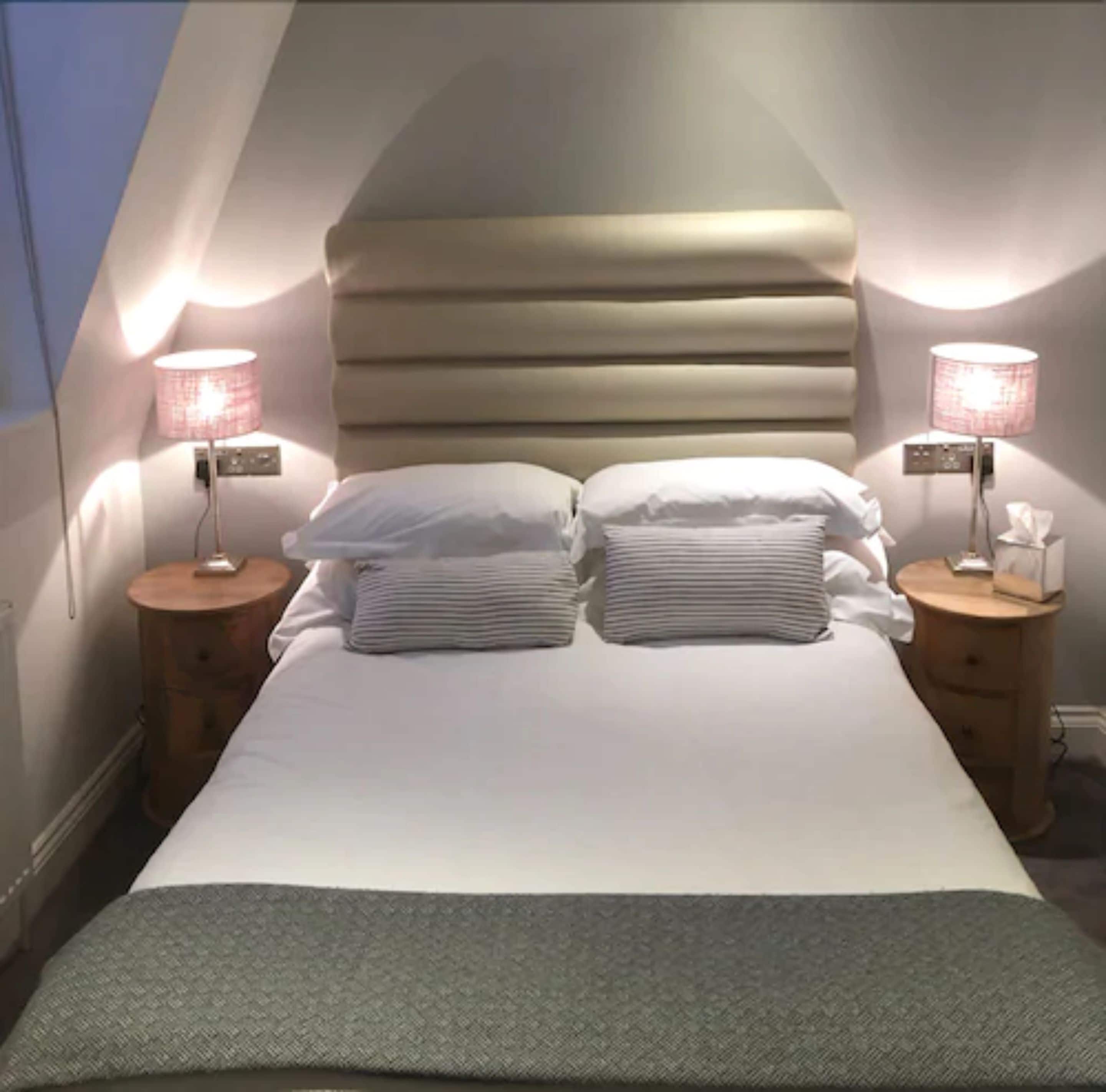 Ein Schlafzimmer mit schrägem Dach, einem Bett mit weißer Bettwäsche, zwei Nachttischen mit Lampen und einem Fenster links.