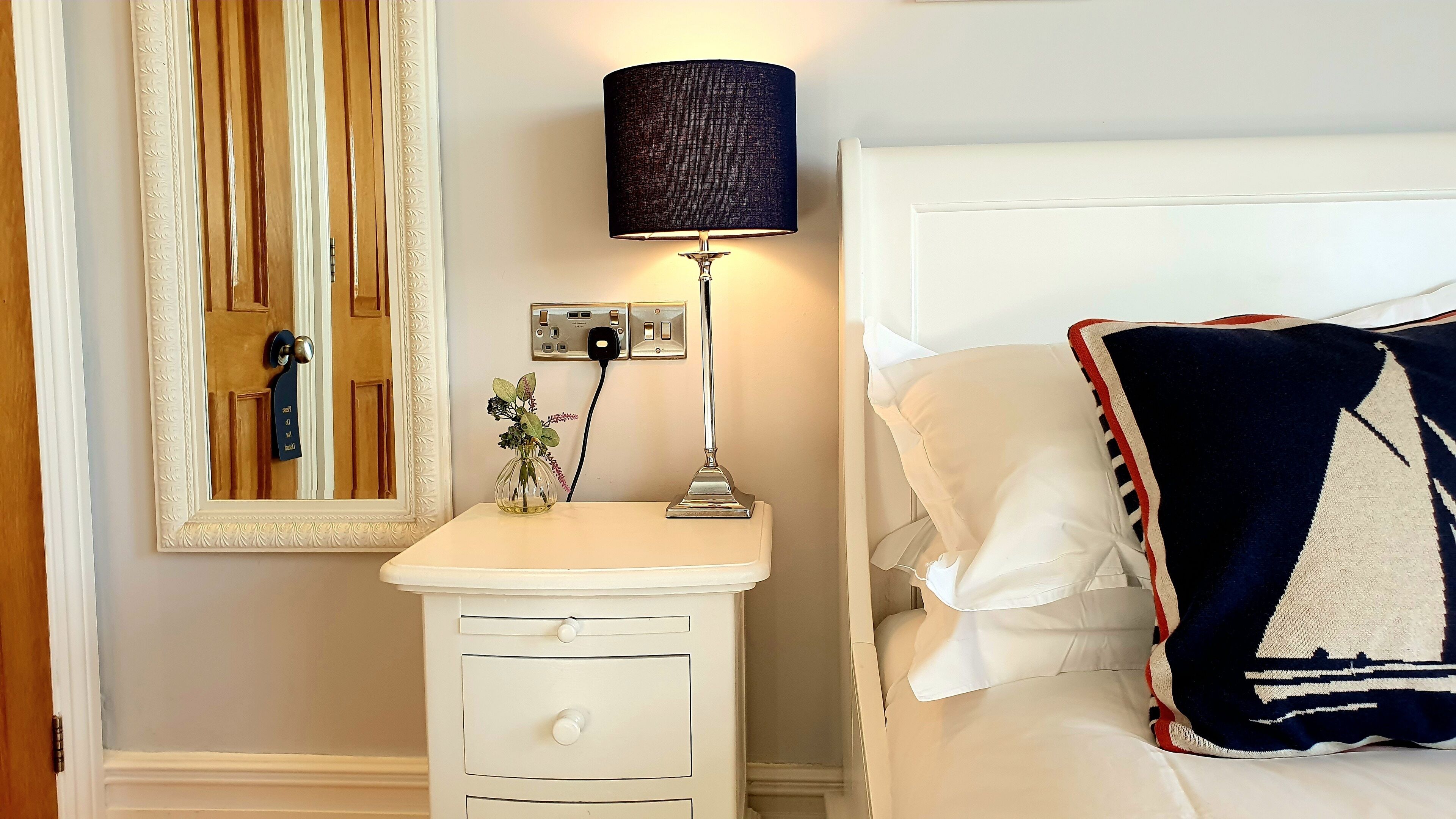 Una habitación de hotel con una cama, una mesita de noche con lámpara, un espejo y un enchufe en la pared.