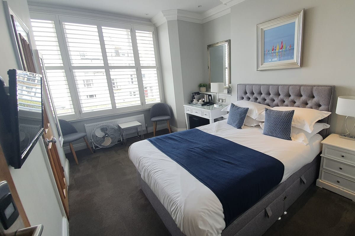 A hotel room with a large bed, a desk, a chair, a mirror, and a window with blinds.
