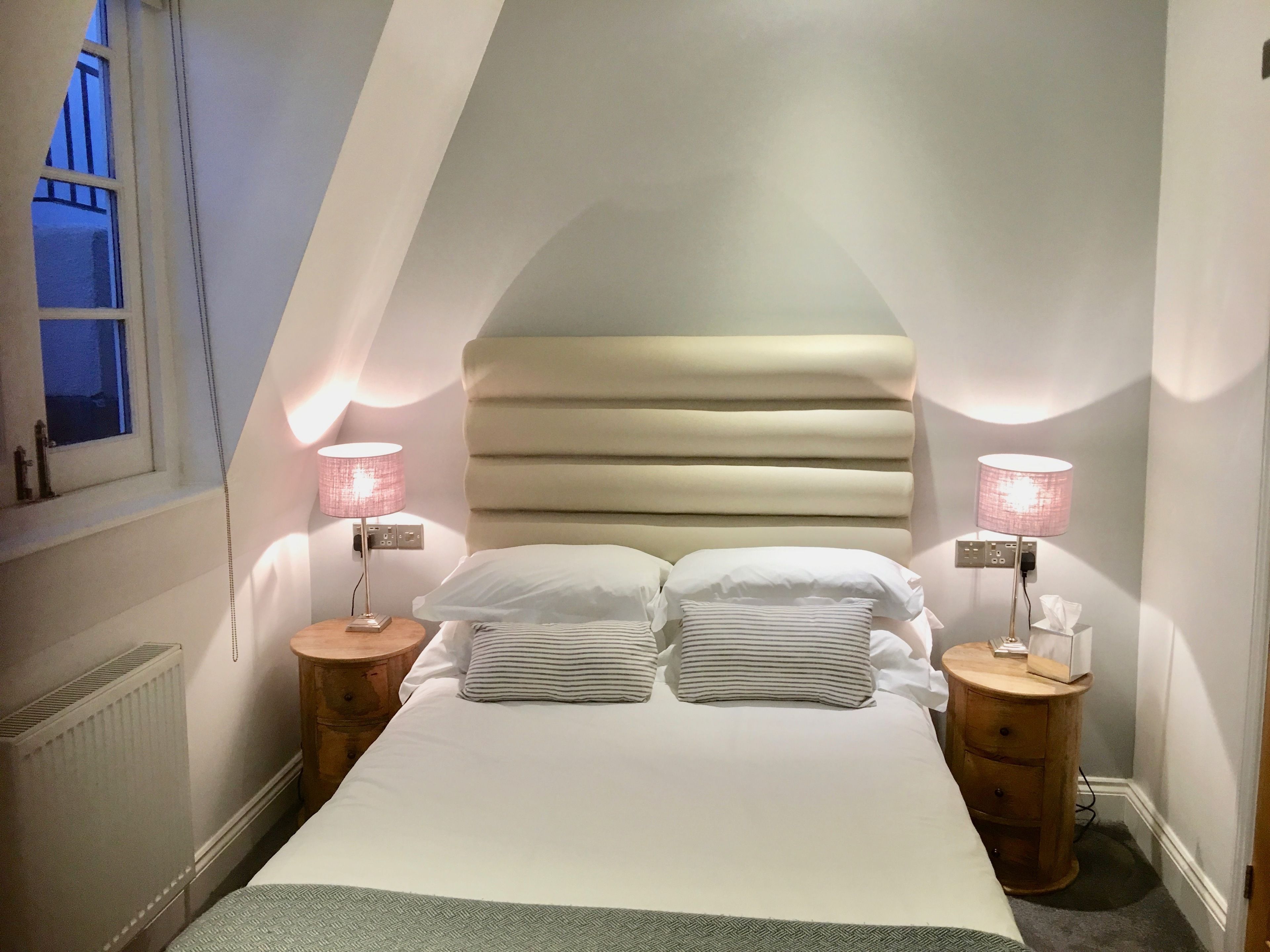 A bedroom with a sloped ceiling, a bed with white bedding, two bedside lamps, and a radiator.