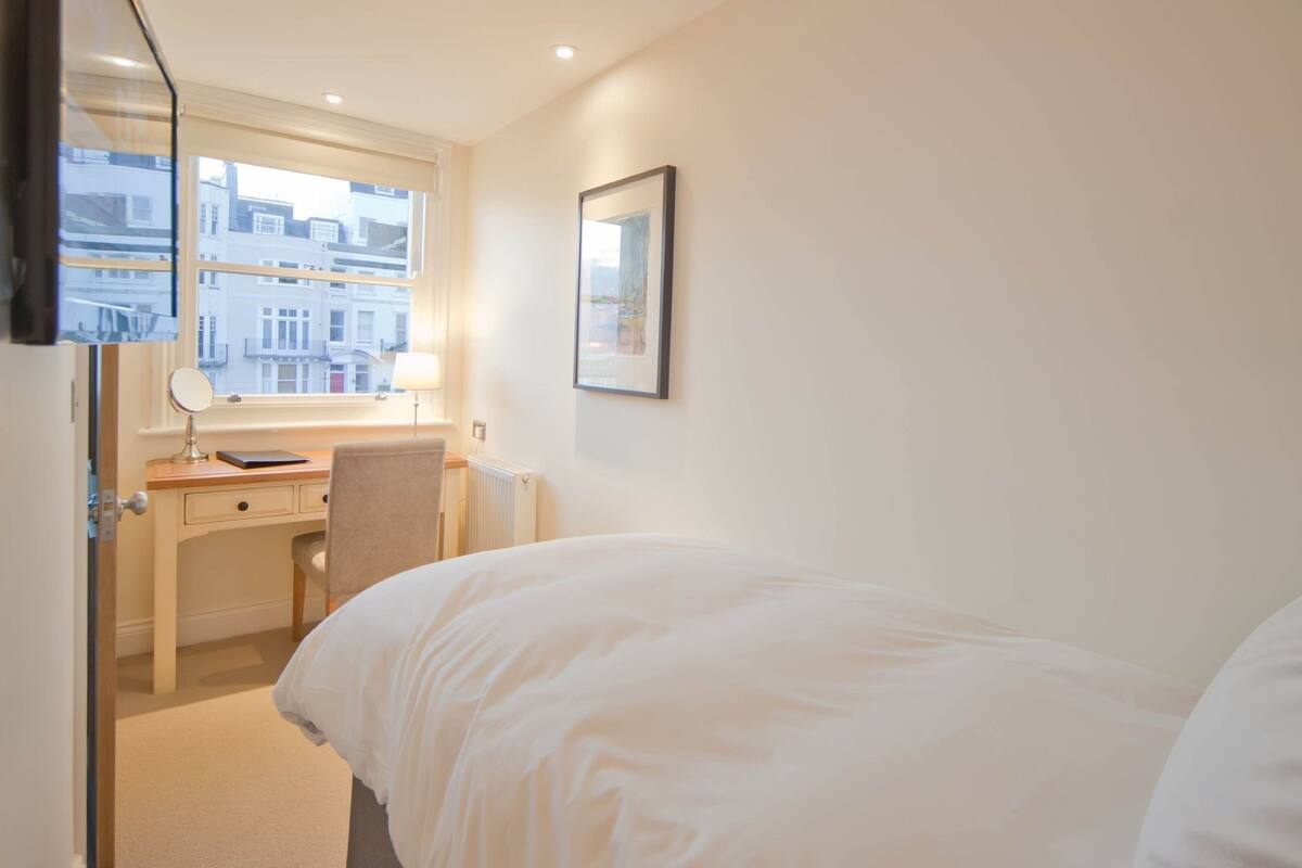 A hotel room with a bed, a desk with a chair, a window with a view of buildings, and a framed picture on the wall.