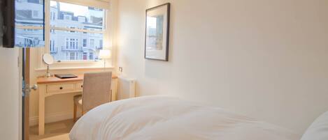 A hotel room with a bed, a desk with a chair, a window with a view of buildings, and a framed picture on the wall.