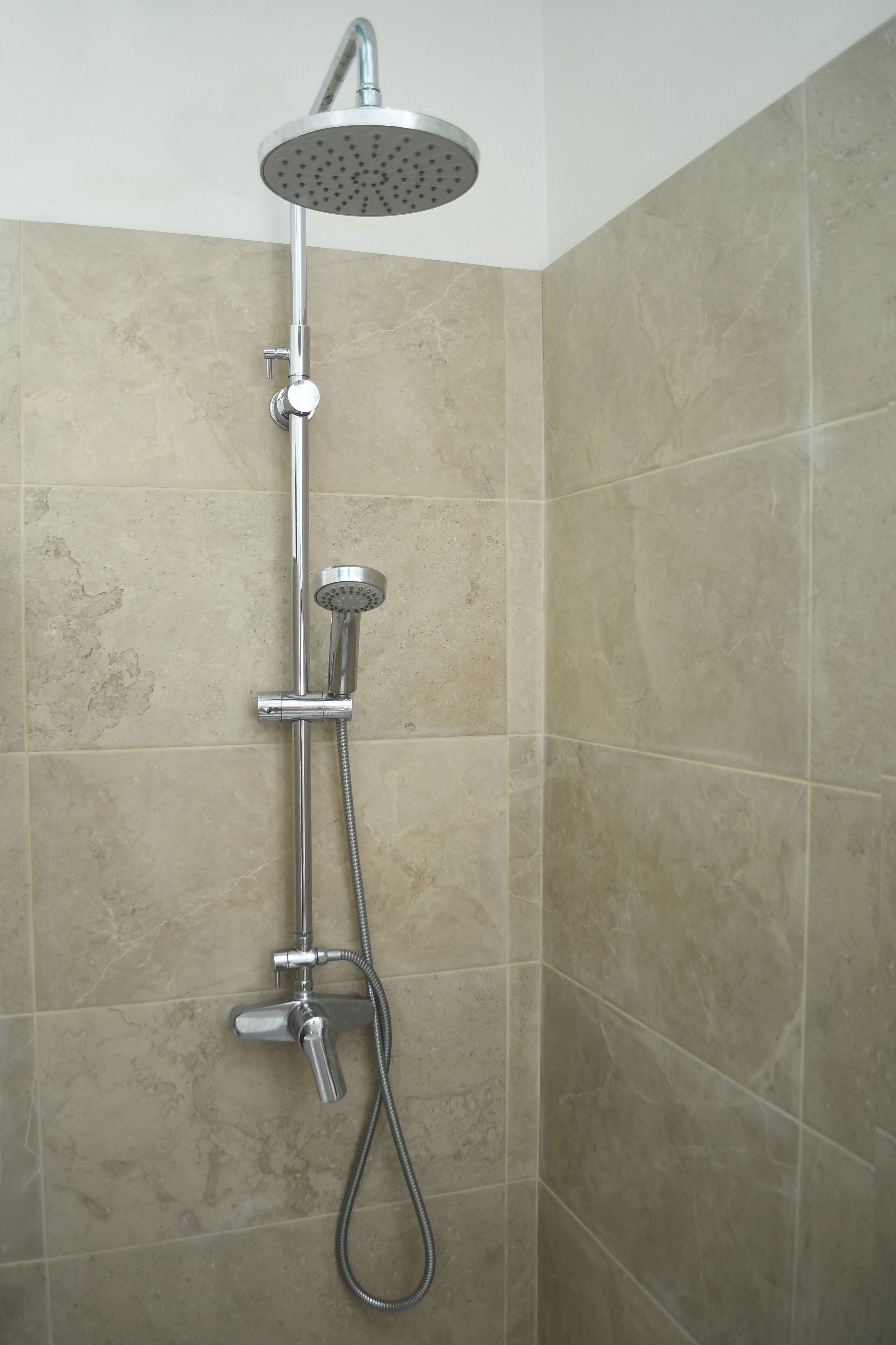 Une douche équipée d’un pommeau de douche à main et d’un pommeau de douche fixe au plafond, le tout installé sur un mur recouvert de carreaux de couleur beige.
