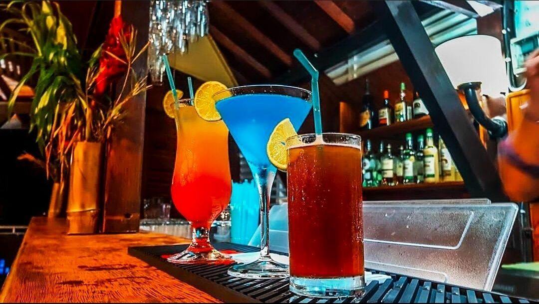 Trois cocktails sont posés sur le comptoir d’un bar, dont la surface est en bois, à côté d’un seau à glace en métal.