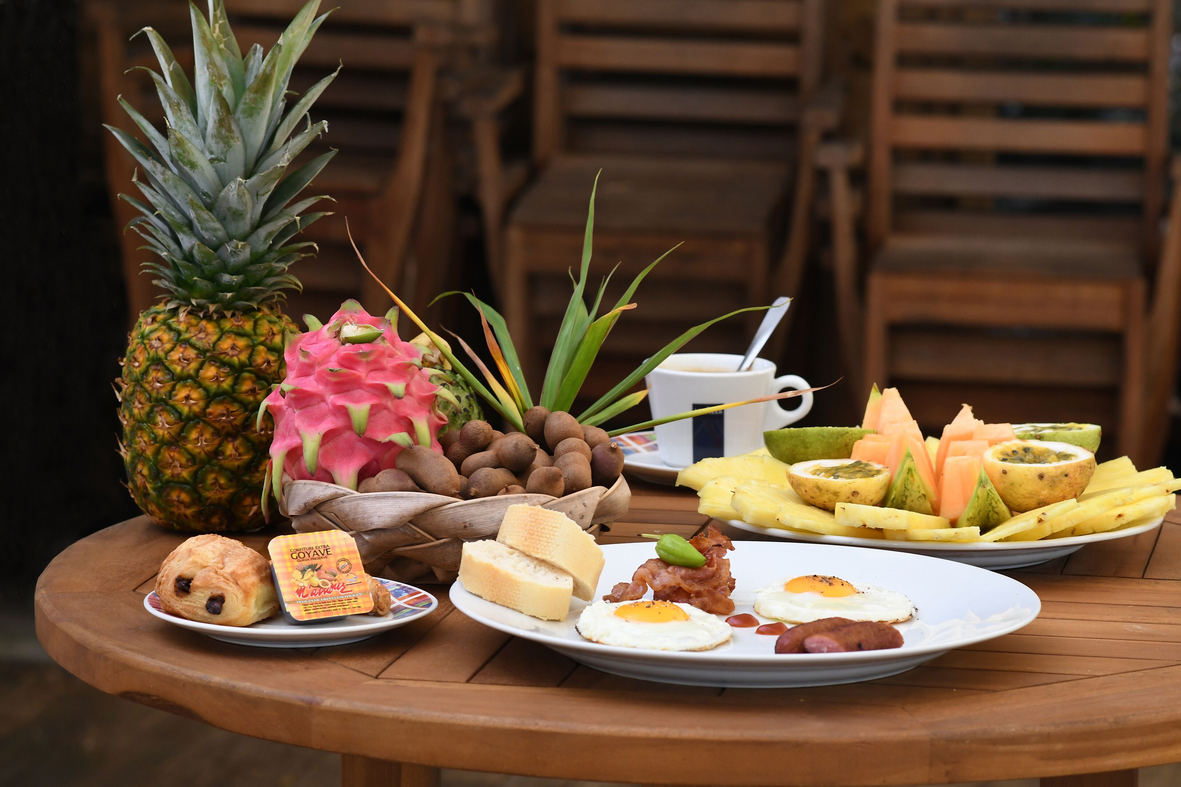 Una mesa de desayuno con diversos alimentos, entre ellos una piña, pitahaya, pan, huevos y un plato de fruta.