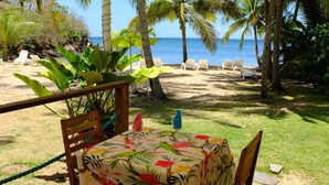 Une table placée sur la plage, recouverte d’une nappe à motifs floraux, entourée de chaises, et offrant une vue sur l’océan et les palmiers.