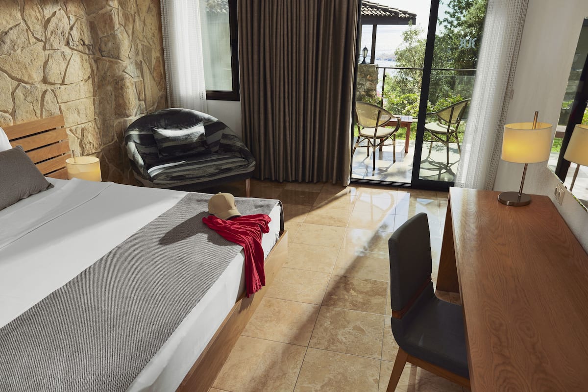A hotel room with a bed, a desk, a chair, a red throw blanket, and a view of the outdoors through a glass door.