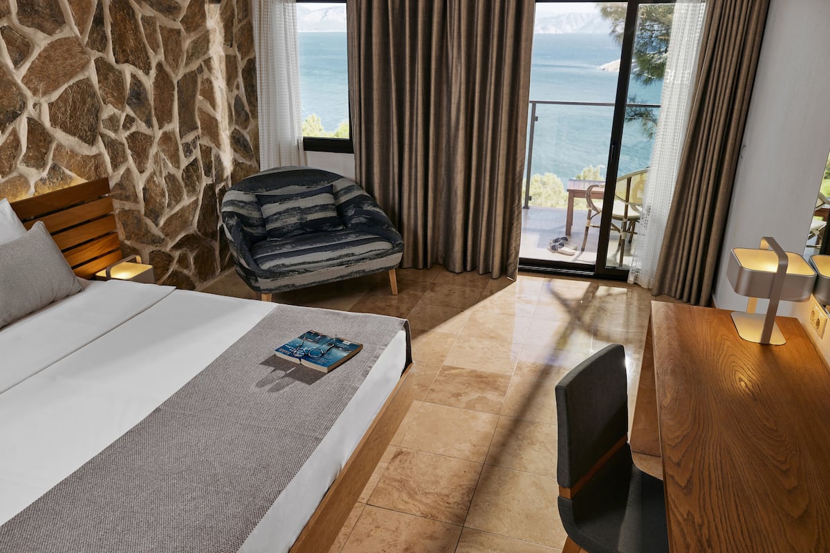 A bedroom with a stone wall, a bed, a chair, a desk, and a view of the sea.