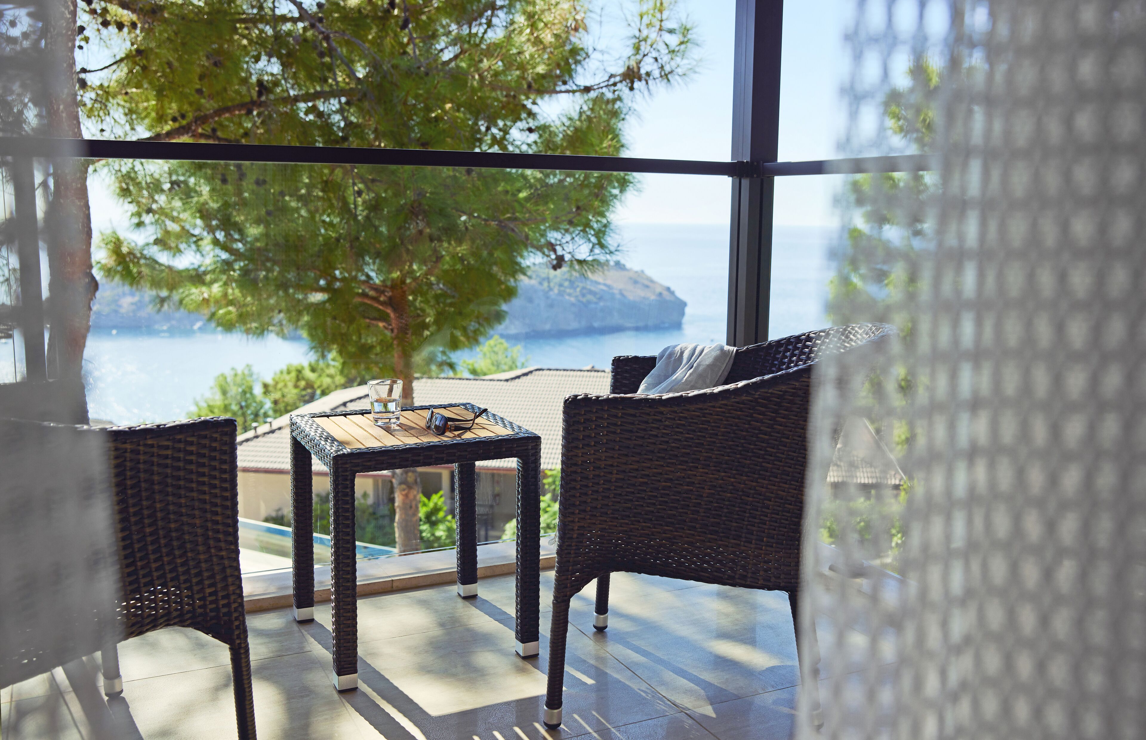 Uma varanda com móveis de vime, uma mesa e vista para o mar e uma árvore.