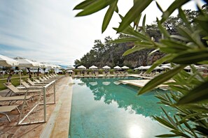 Une piscine entourée de chaises longues et de parasols, bordée de verdure et adossée à une colline rocheuse.