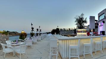 A rooftop bar with white wicker chairs and tables, a bar counter with bottles, and a view of the sea.