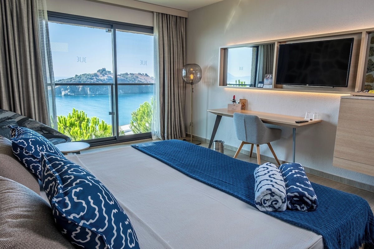 A bedroom with a large bed, a desk, a TV, and a view of the sea.