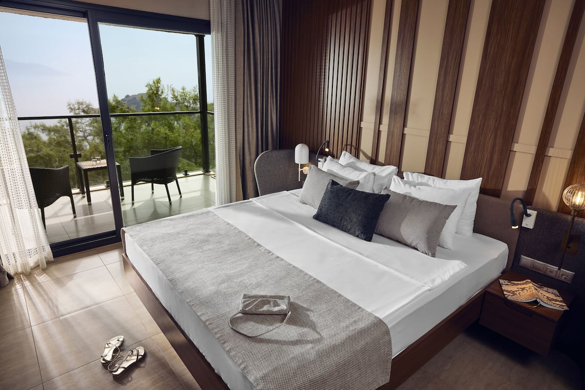 A modern hotel room with a large bed, a balcony with a view, and a nightstand with a lamp.