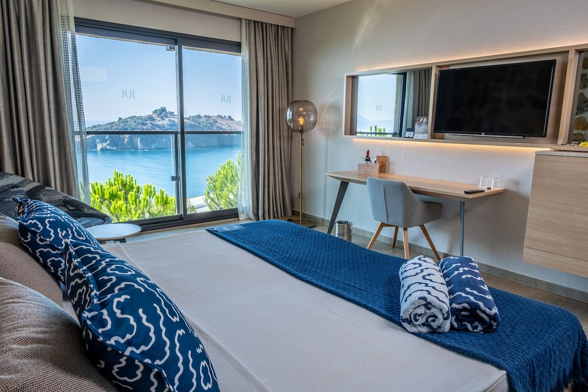 A hotel room with a bed, a desk, a TV, and a view of the sea.