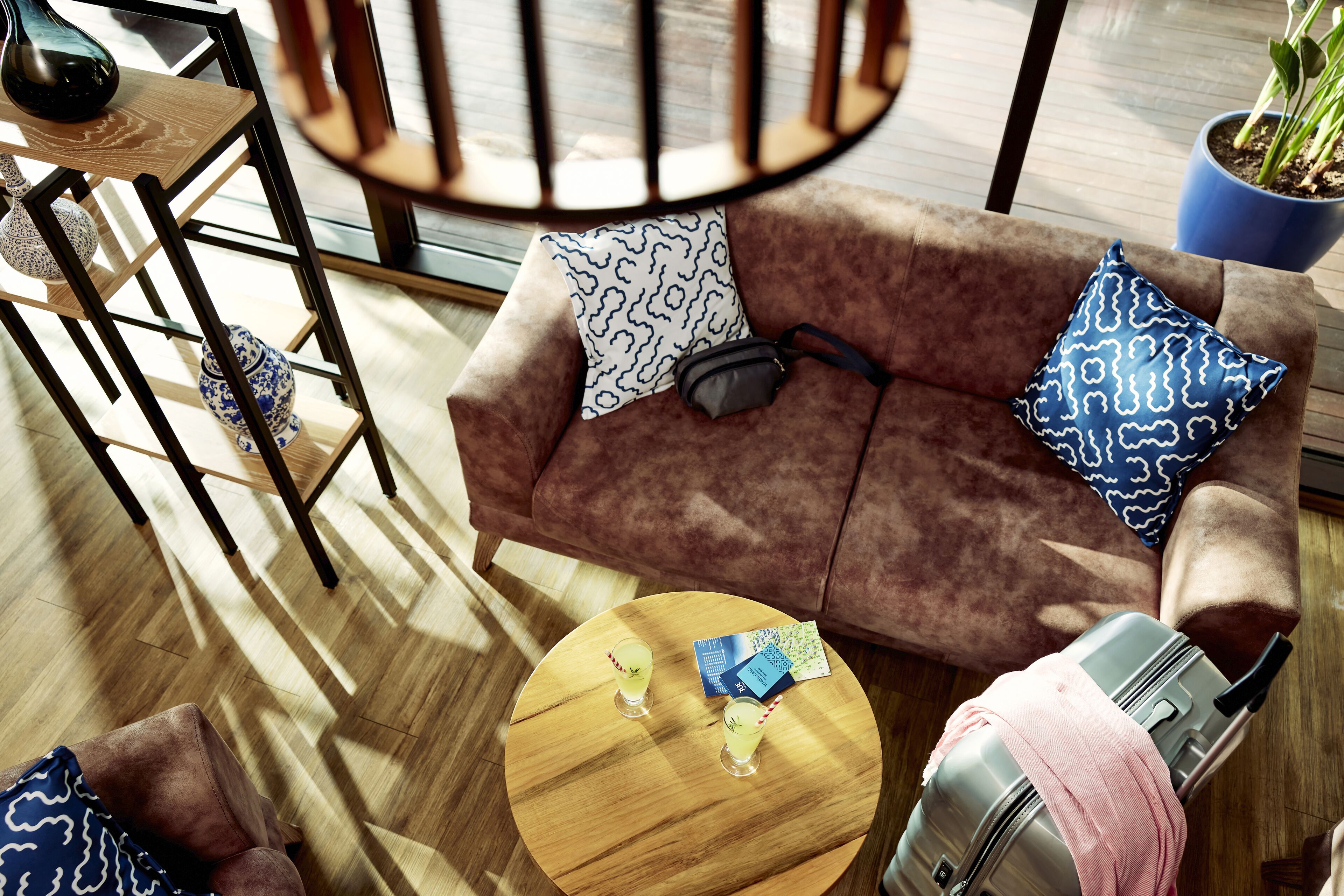 Un salon moderne avec un canapé marron, des coussins à motifs, une petite table ronde sur laquelle sont posées des boissons et une valise.