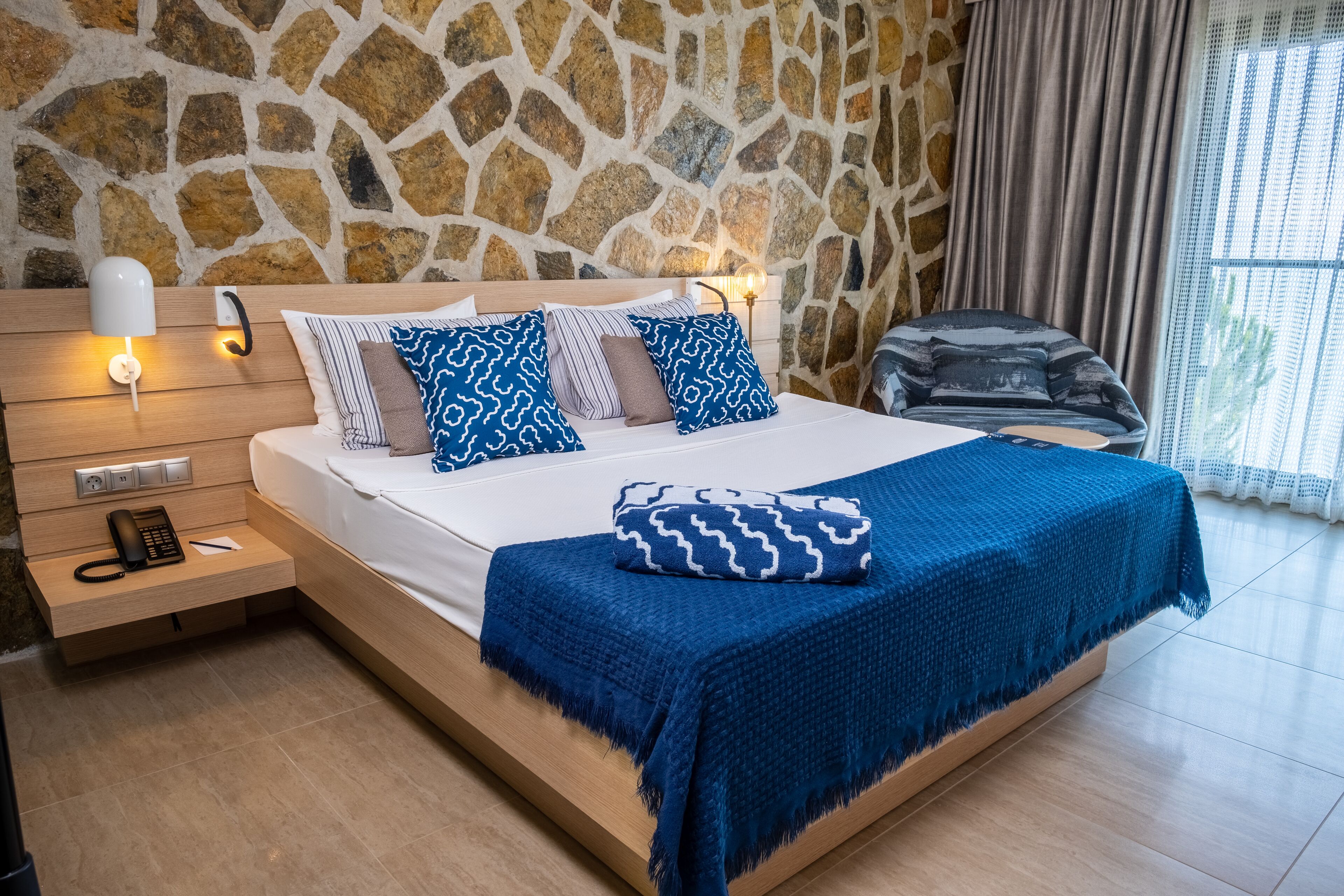 A bedroom with a stone wall, a bed with blue and white patterned pillows, a blue blanket, bedside lamps, and a wooden nightstand with a phone.