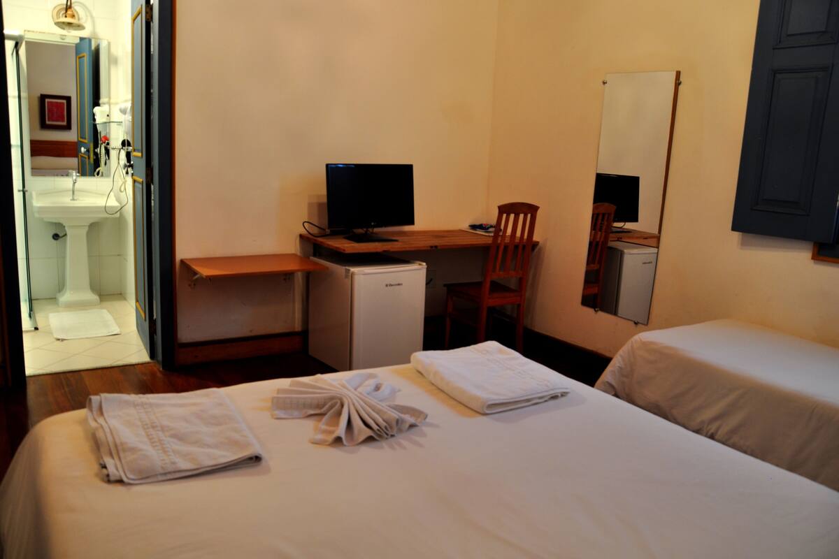Hotelkamer met een bed, een bureau met een computer, een stoel, een kleine koelkast en een badkamer zichtbaar door een geopende deur.