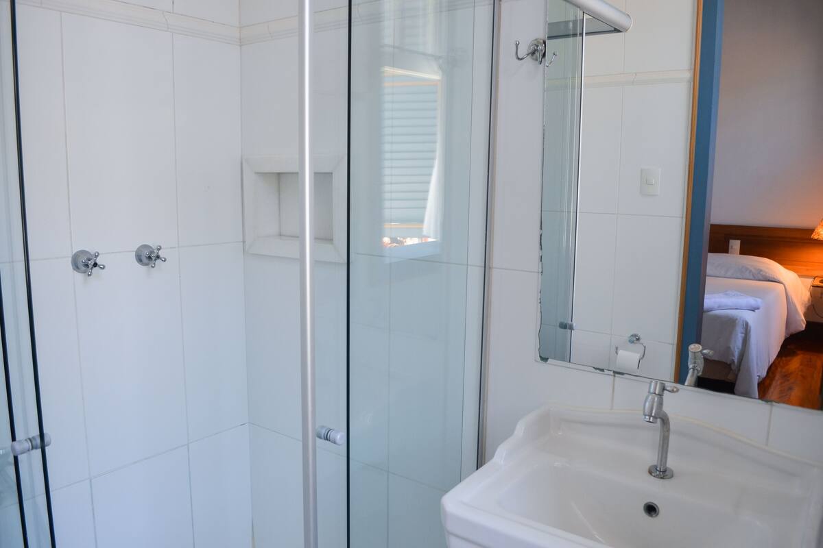 Een moderne badkamer met een douchecabine van glas, een witte wastafel en een spiegel die een bed en raam weerkaatst.
