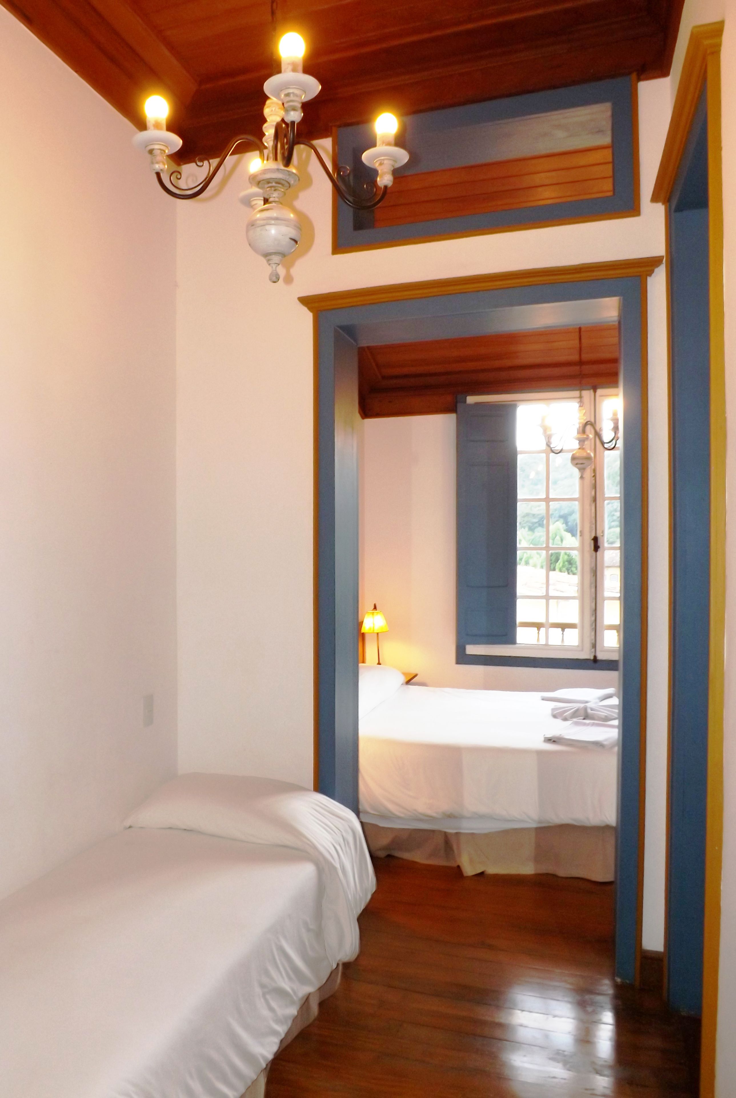 A bedroom with a wooden ceiling, a chandelier, a bed, and a window with blue shutters.