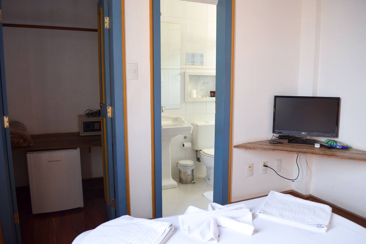 Hotelkamer met een bed, een bureau met een computer, een televisie, een kleine koelkast en een badkamer zichtbaar door een geopende deur.