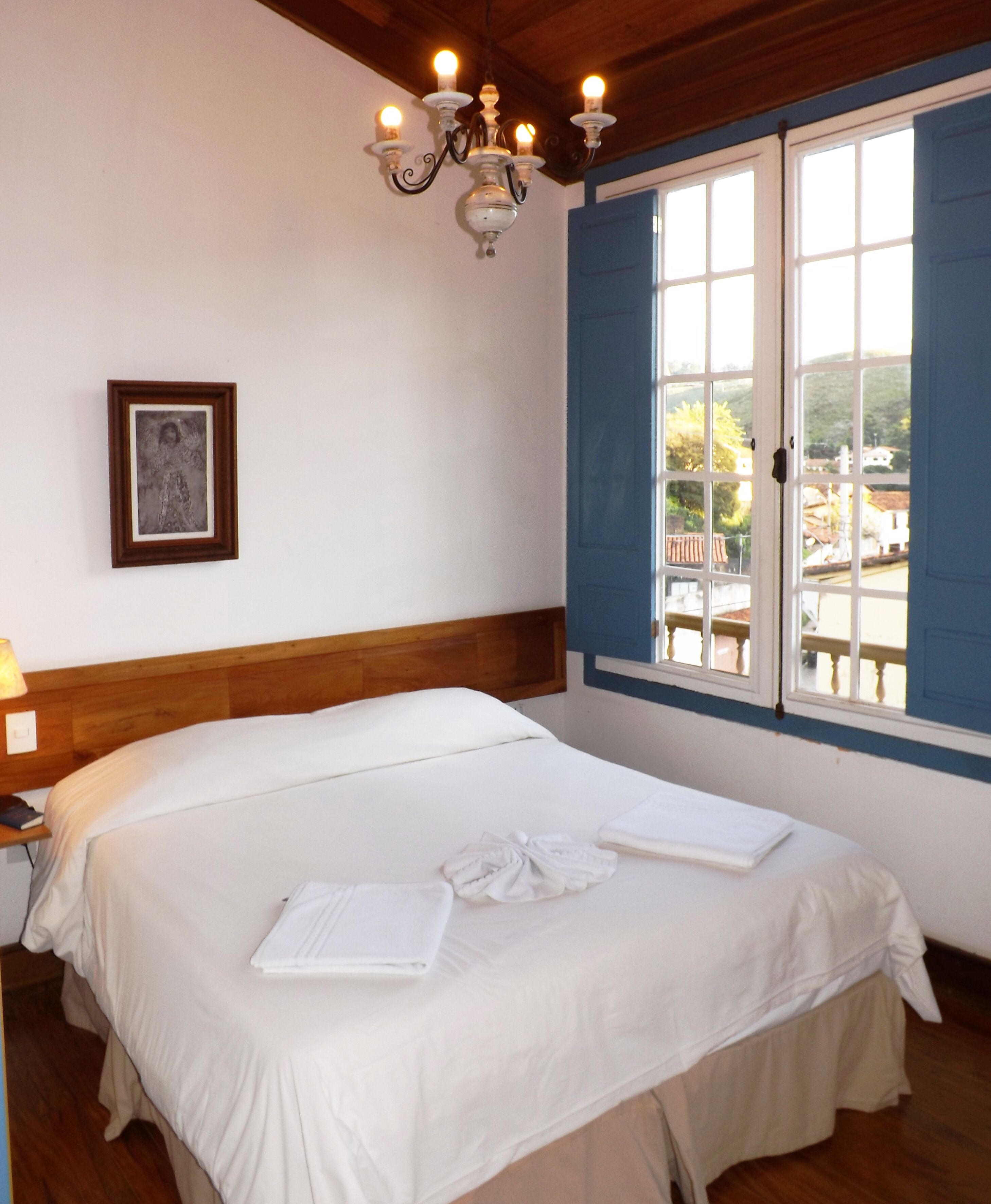 Una habitación de hotel con una cama grande, una araña, un cuadro en la pared y una ventana con contraventanas azules.