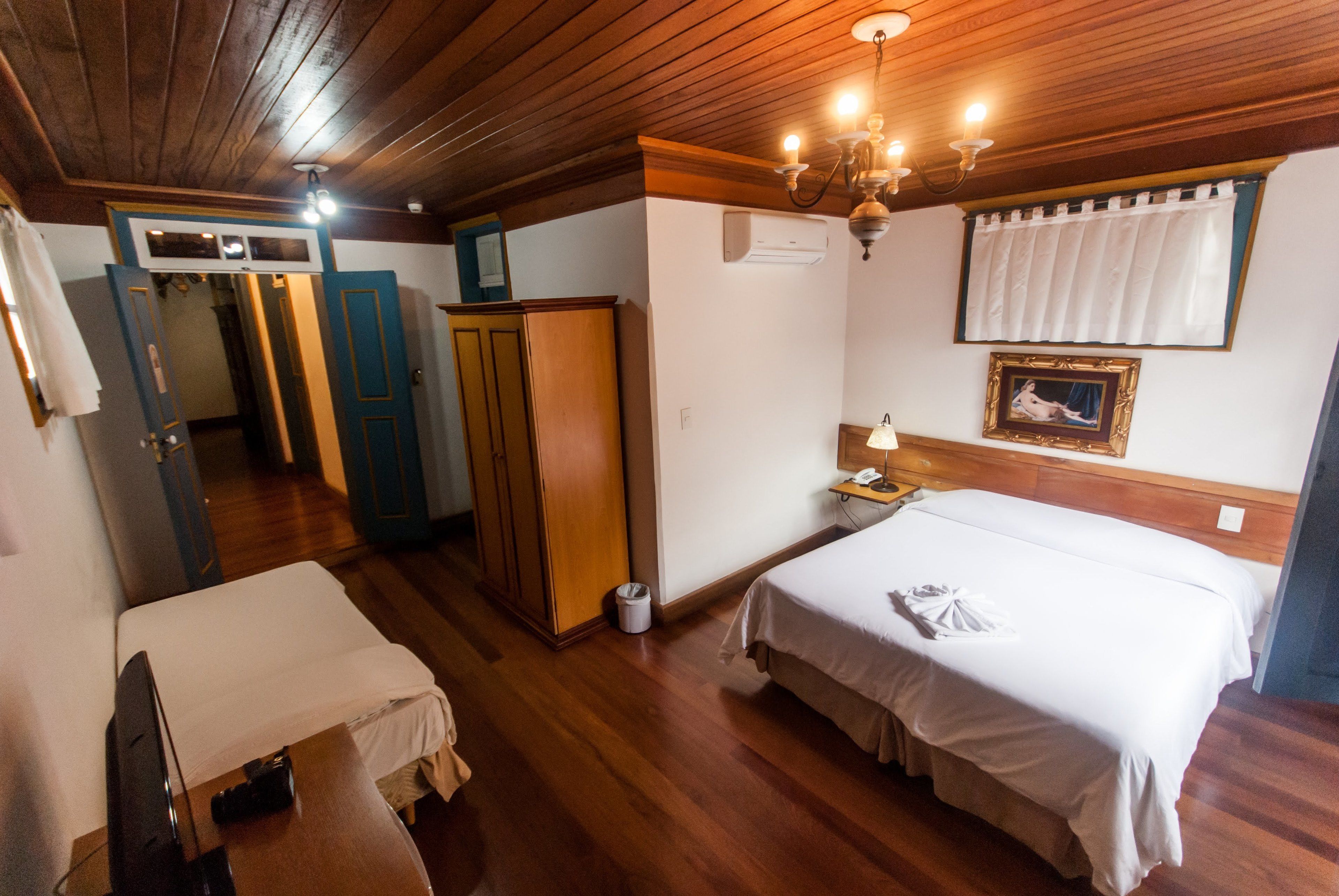 A hotel room with two beds, wooden flooring, a wardrobe, a desk with a lamp, and a chandelier.