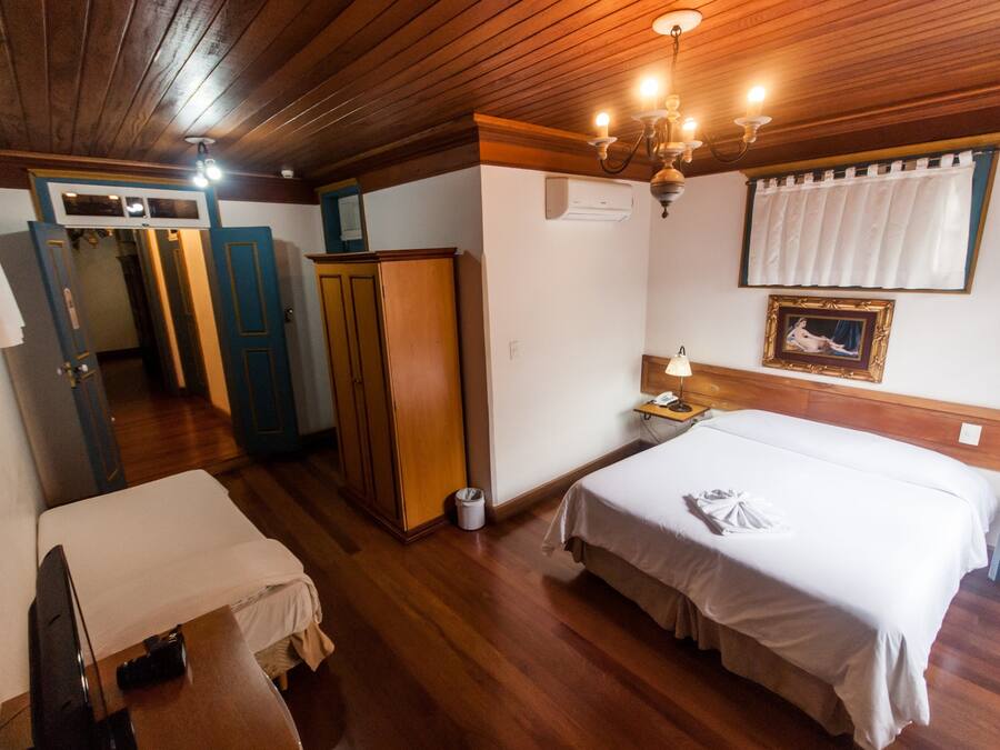 A hotel room with two beds, wooden flooring, a wardrobe, a desk with a lamp, and a chandelier.