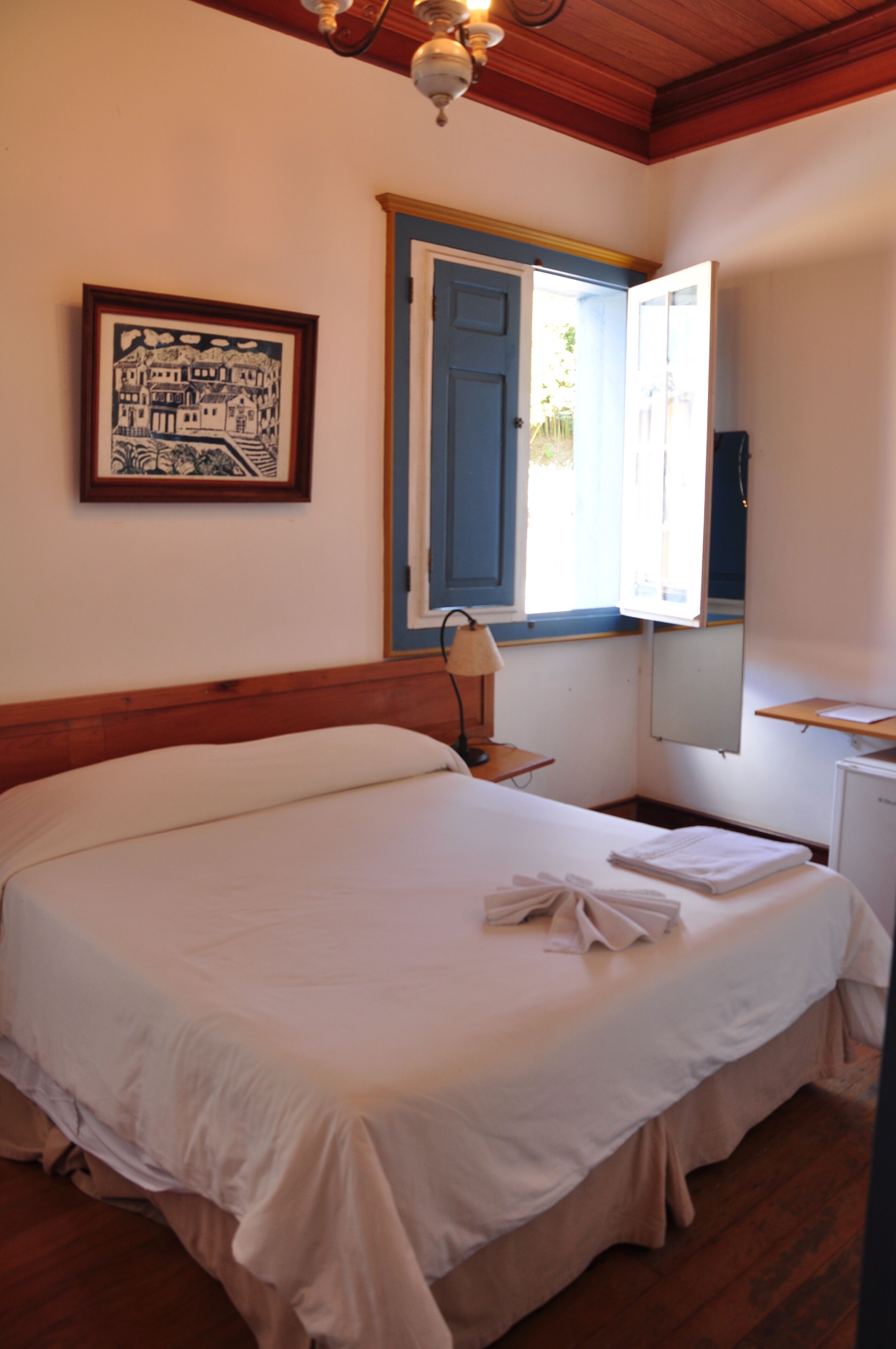A bedroom with a bed, a nightstand, a lamp, a window with an open door, a framed picture on the wall, and a small desk with a mirror.