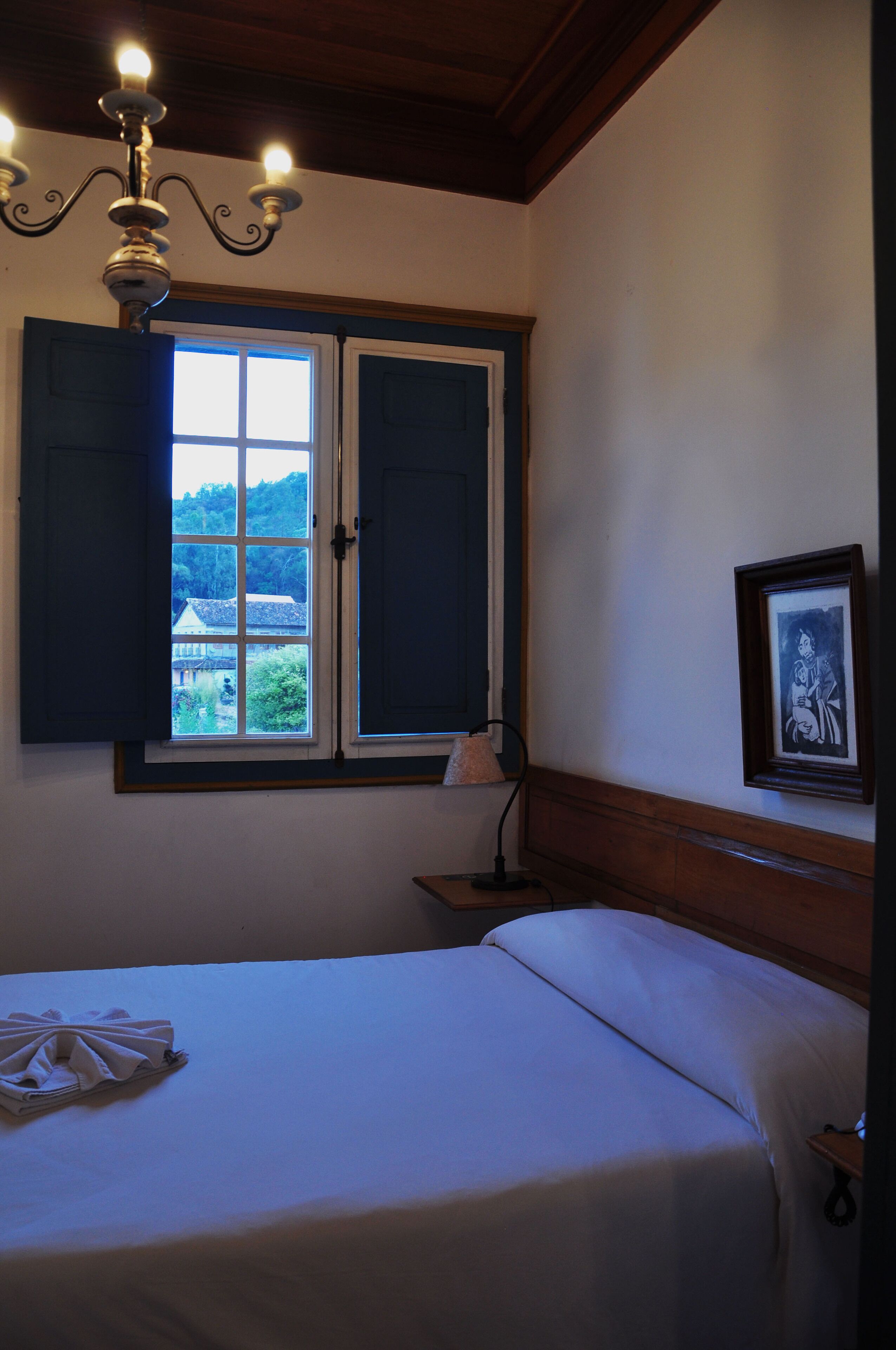 Un dormitorio con cama, ventana con contraventanas, candelabro, mesita de noche y un cuadro en la pared.