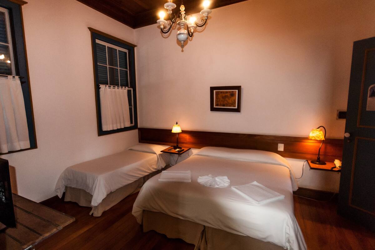 Een hotelkamer met twee eenpersoonsbedden, een houten plafond, een kroonluchter, een raam met gordijnen en een nachtkastje met een lamp.