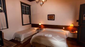 A hotel room with two single beds, a wooden ceiling, a chandelier, a window with curtains, and a bedside table with a lamp.