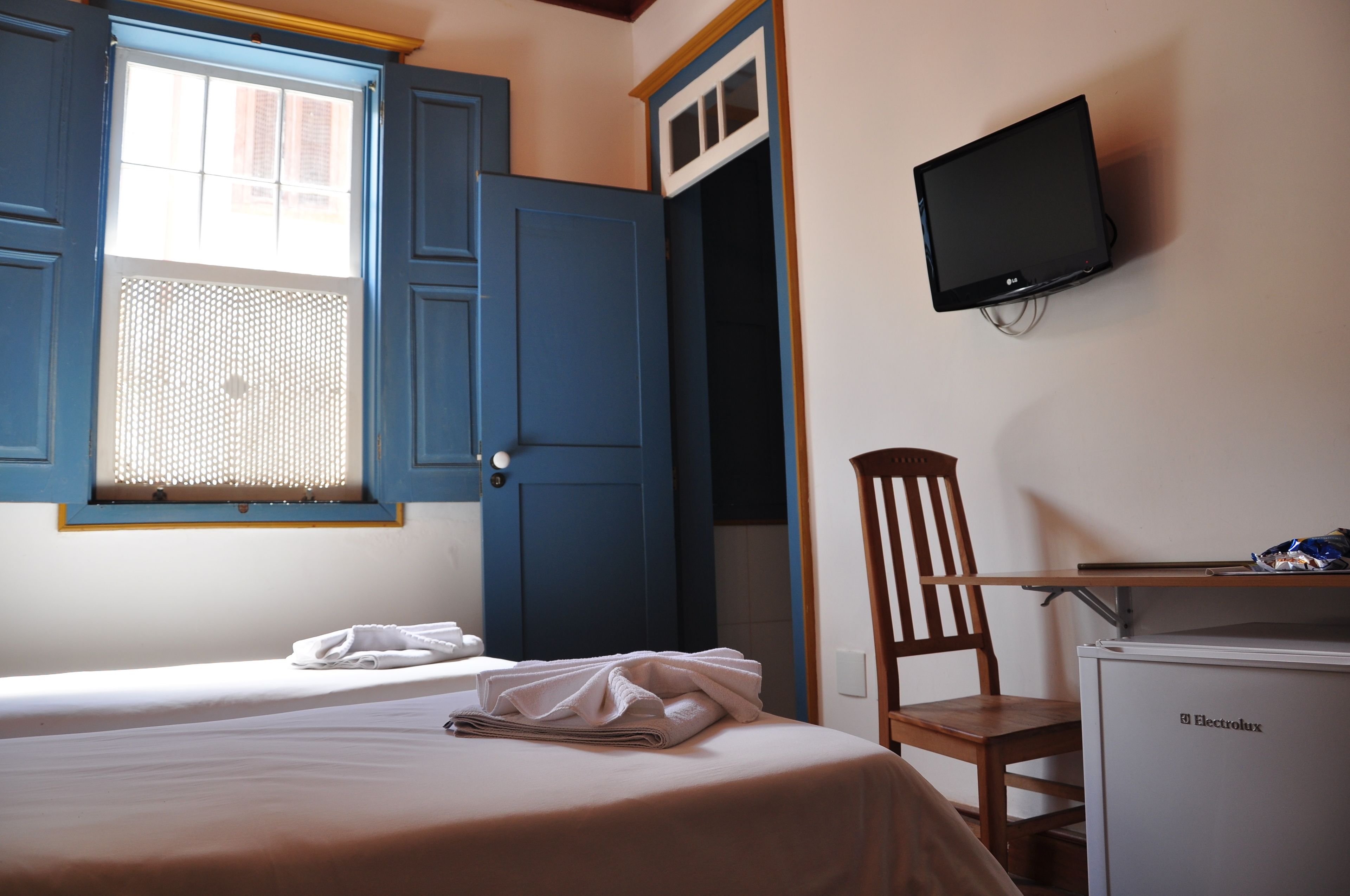Een slaapkamer met een bed, een stoel, een bureau, een televisie en een raam met blauwe luiken.