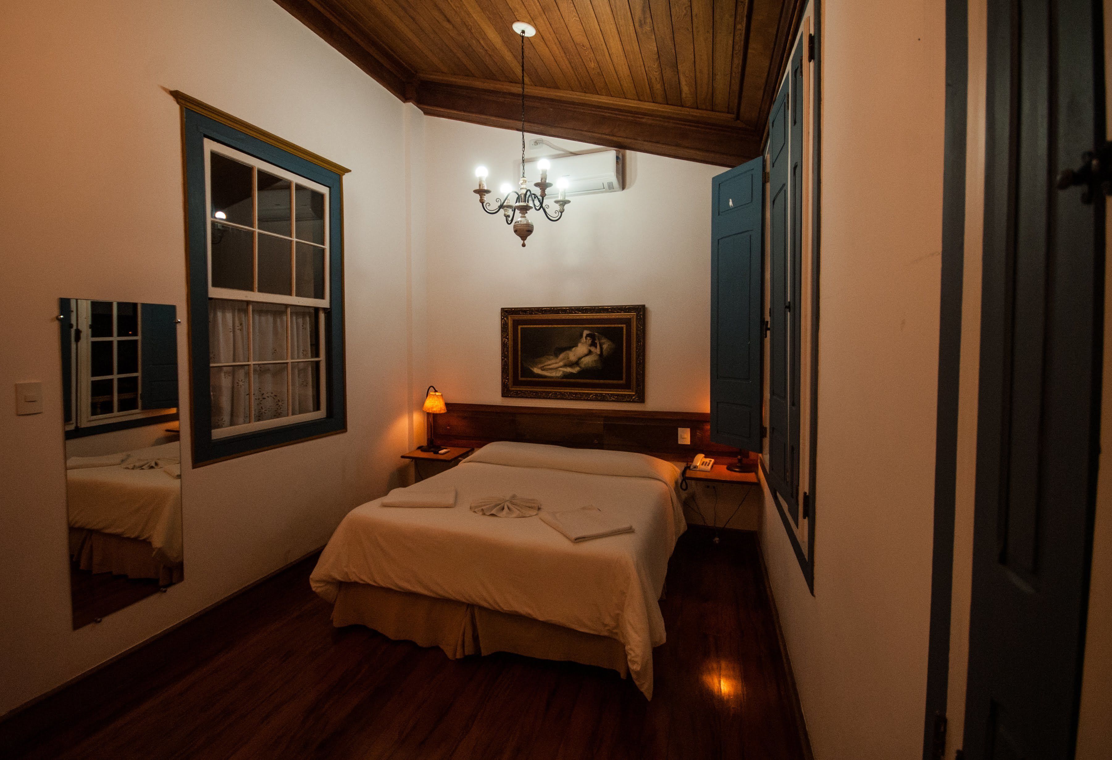 A bedroom with a wooden ceiling, a bed with a white bedspread, a chandelier, a framed picture on the wall, and a window with curtains.