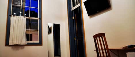 Un dormitorio con cama, escritorio, silla, televisor montado en la pared y ventana con cortinas.