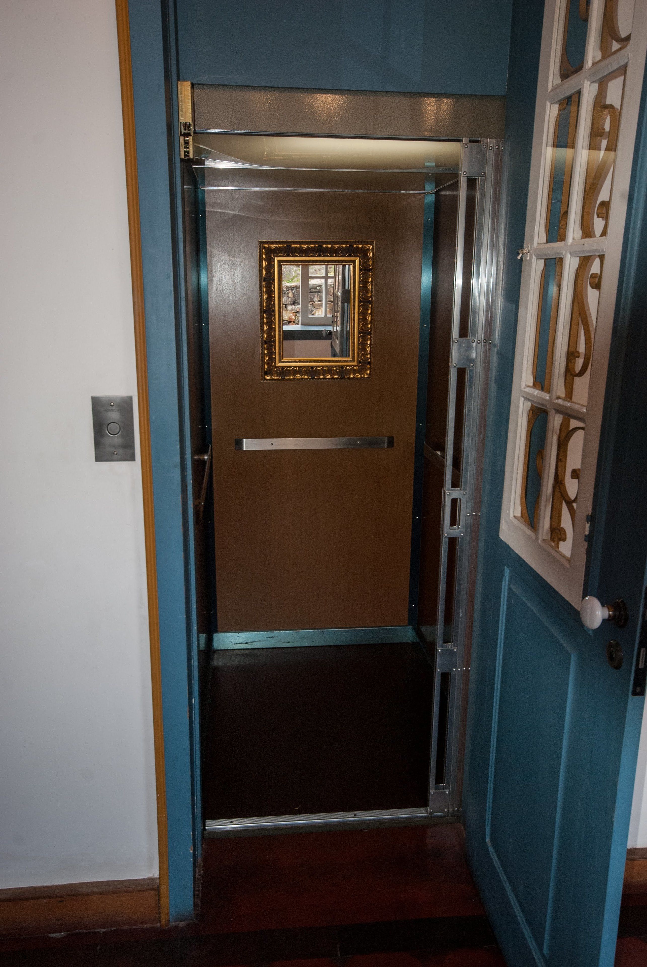 Un ascenseur avec une porte en bois et un cadre en métal, une petite photo encadrée sur la porte, et un mur bleu agrémenté d’une fenêtre décorative.