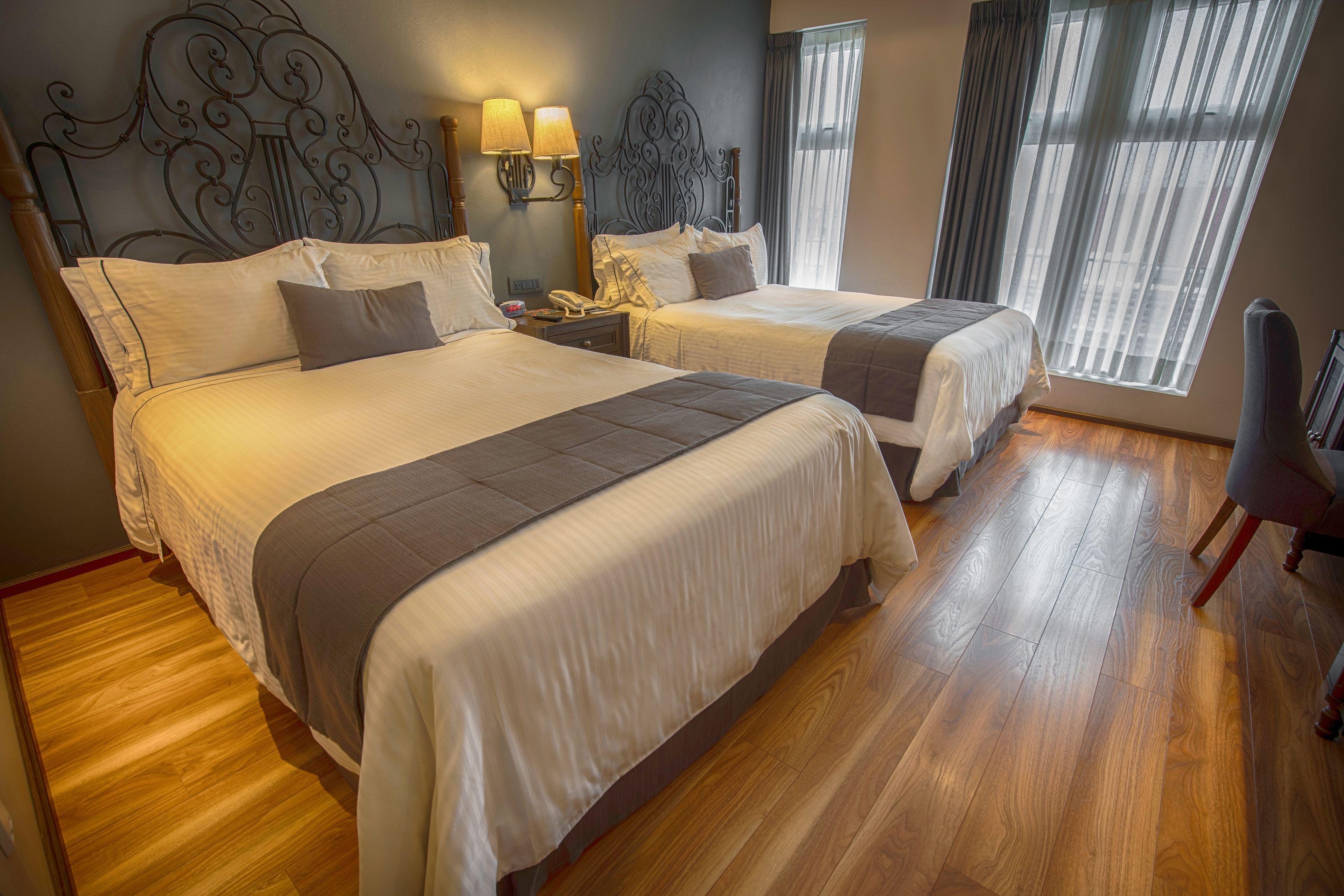 Quarto de hotel com duas camas, piso de madeira, painel decorativo na cabeceira e janela com cortinas.