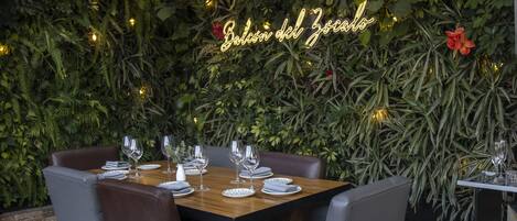 An outdoor dining area with a table set for two, surrounded by lush greenery and a neon sign reading "Balcon del Zocalo".