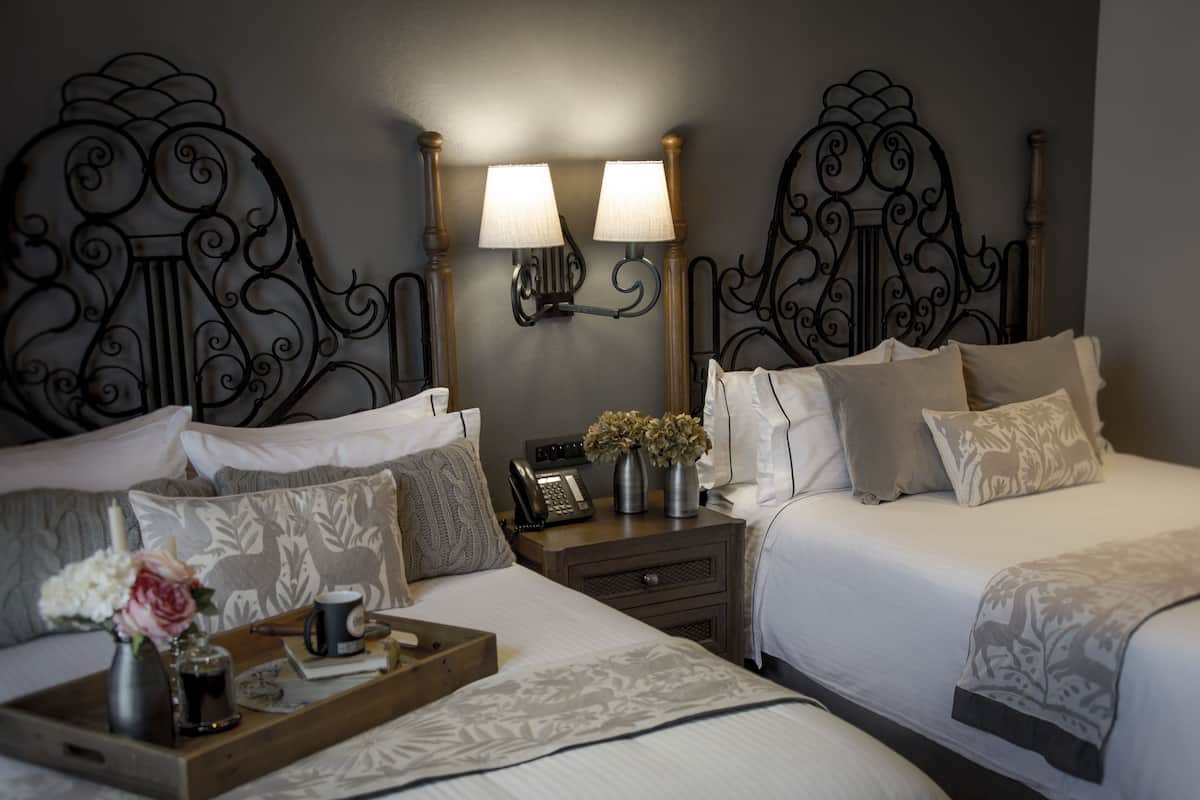 A hotel room with two beds, a bedside table with a lamp, a telephone, and a tray with a vase of flowers.