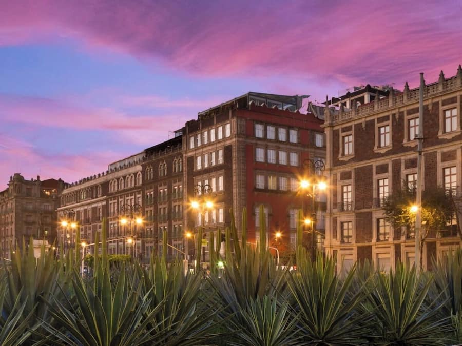 A row of historic buildings with uniform facades and multiple windows, lined with street lamps and surrounded by agave plants.