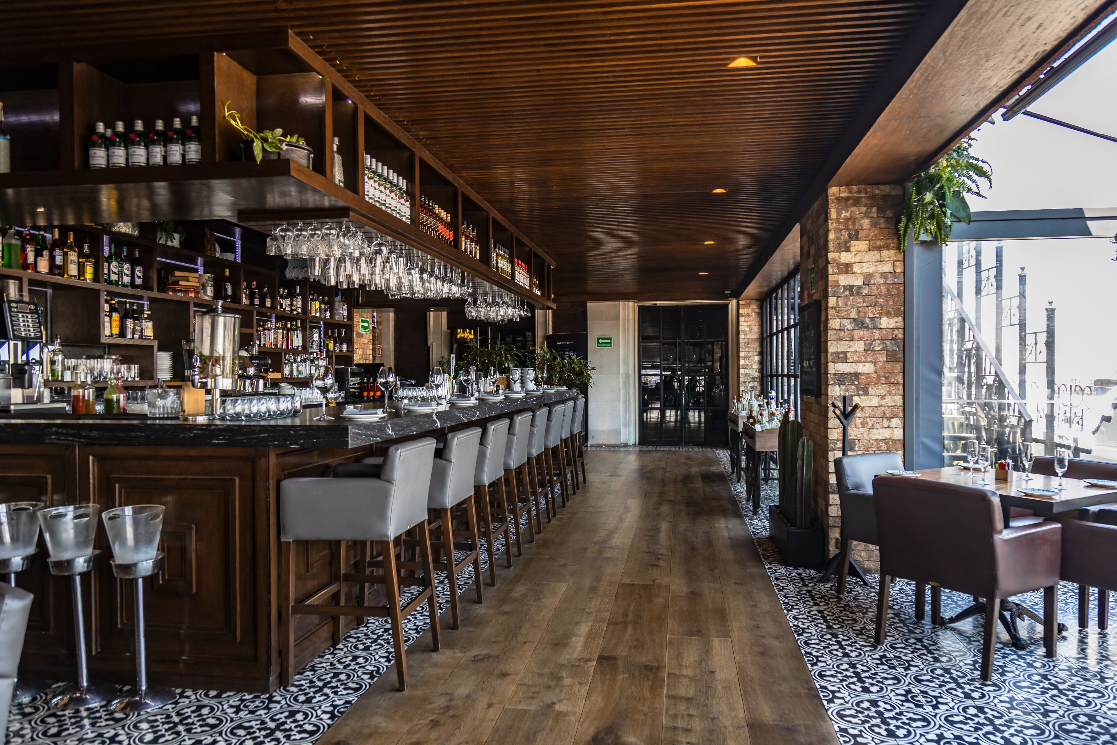 Zona de bar con una barra larga, varios taburetes y una variedad de botellas y vasos.