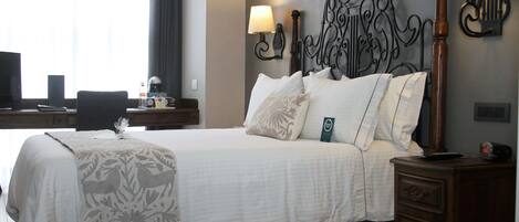 Habitación de hotel con una cama grande, dos lámparas de noche, un cabecero de madera, un escritorio con una computadora y una lámpara, y una ventana con cortinas.