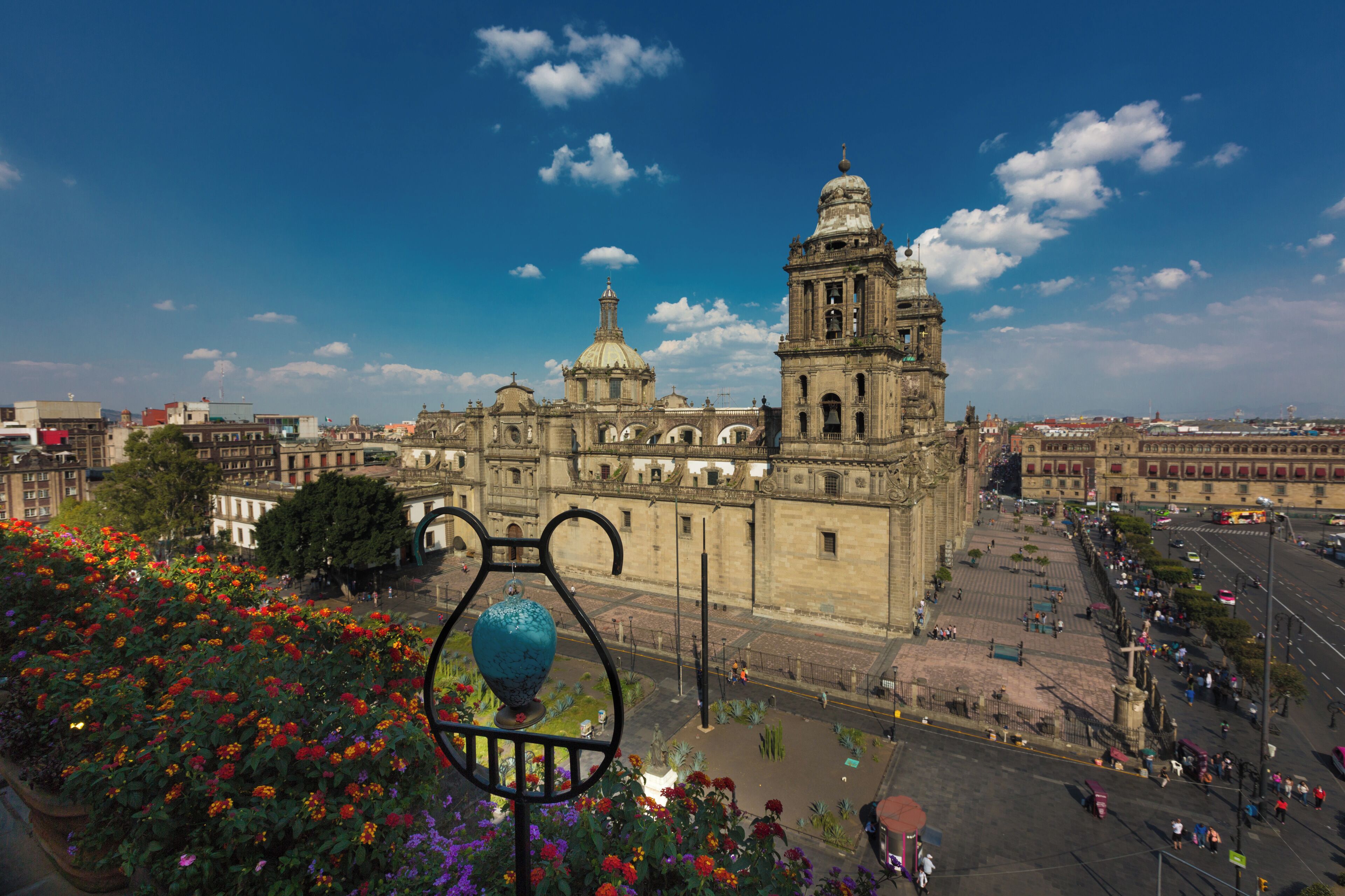 A historic building with a prominent bell tower and dome, surrounded by a plaza with flower beds and a clear blue sky.