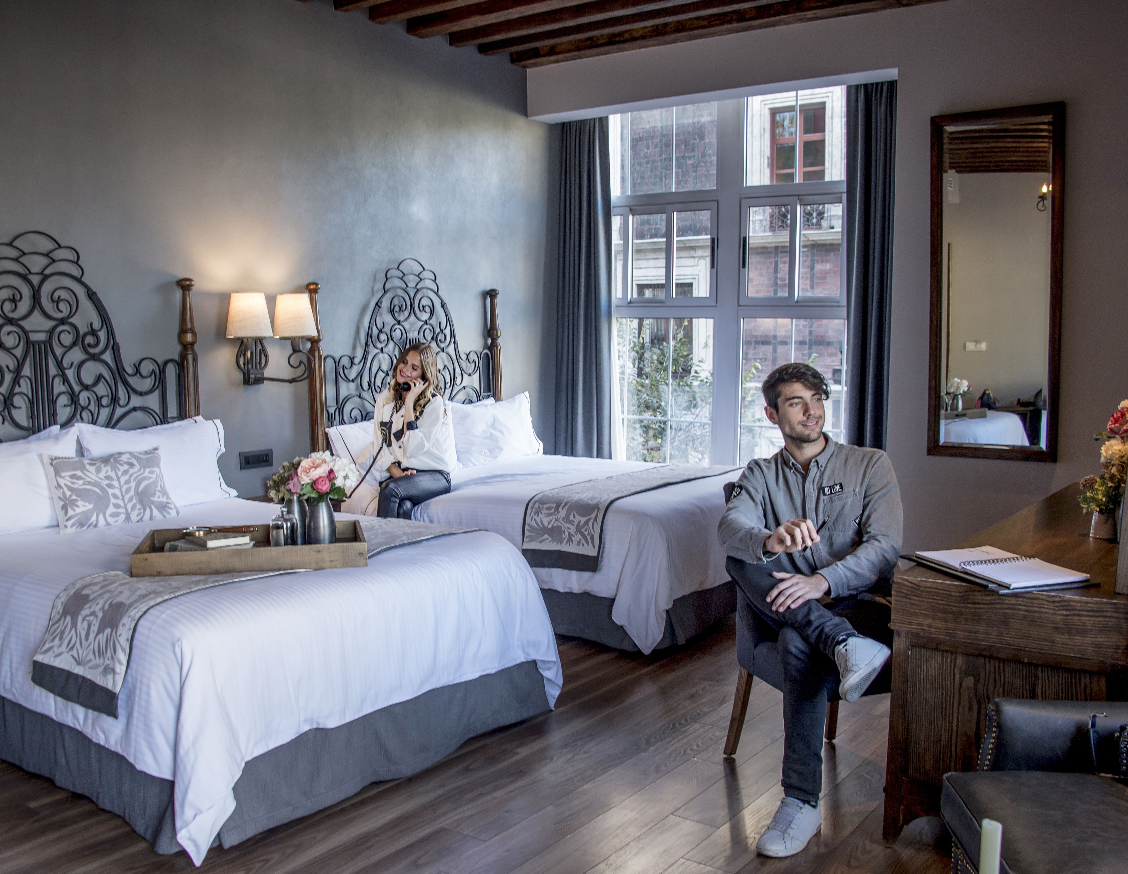 A hotel room with two beds, a wooden floor, a large window, and a mirror on the wall.